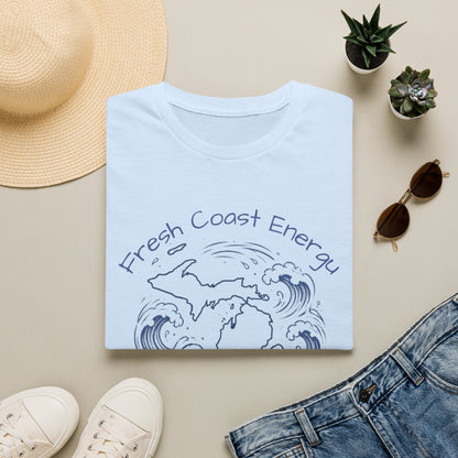 A folded light blue T-shirt with a navy graphic reading 'Fresh Coast Energy' and wave artwork, styled in a flat lay with a straw hat, small potted succulents, sunglasses, white sneakers, and blue jeans on a beige background.