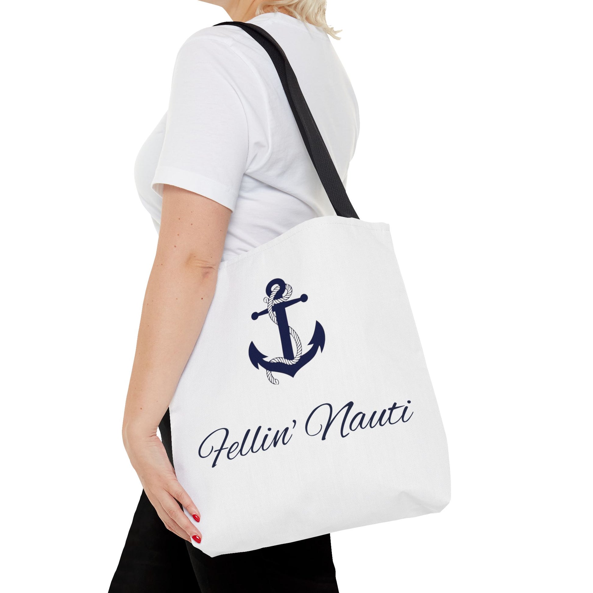 A white canvas tote bag with a black shoulder strap, featuring a navy blue anchor graphic and the text 'Fellin' Nauti' in cursive, shown being worn by a model.