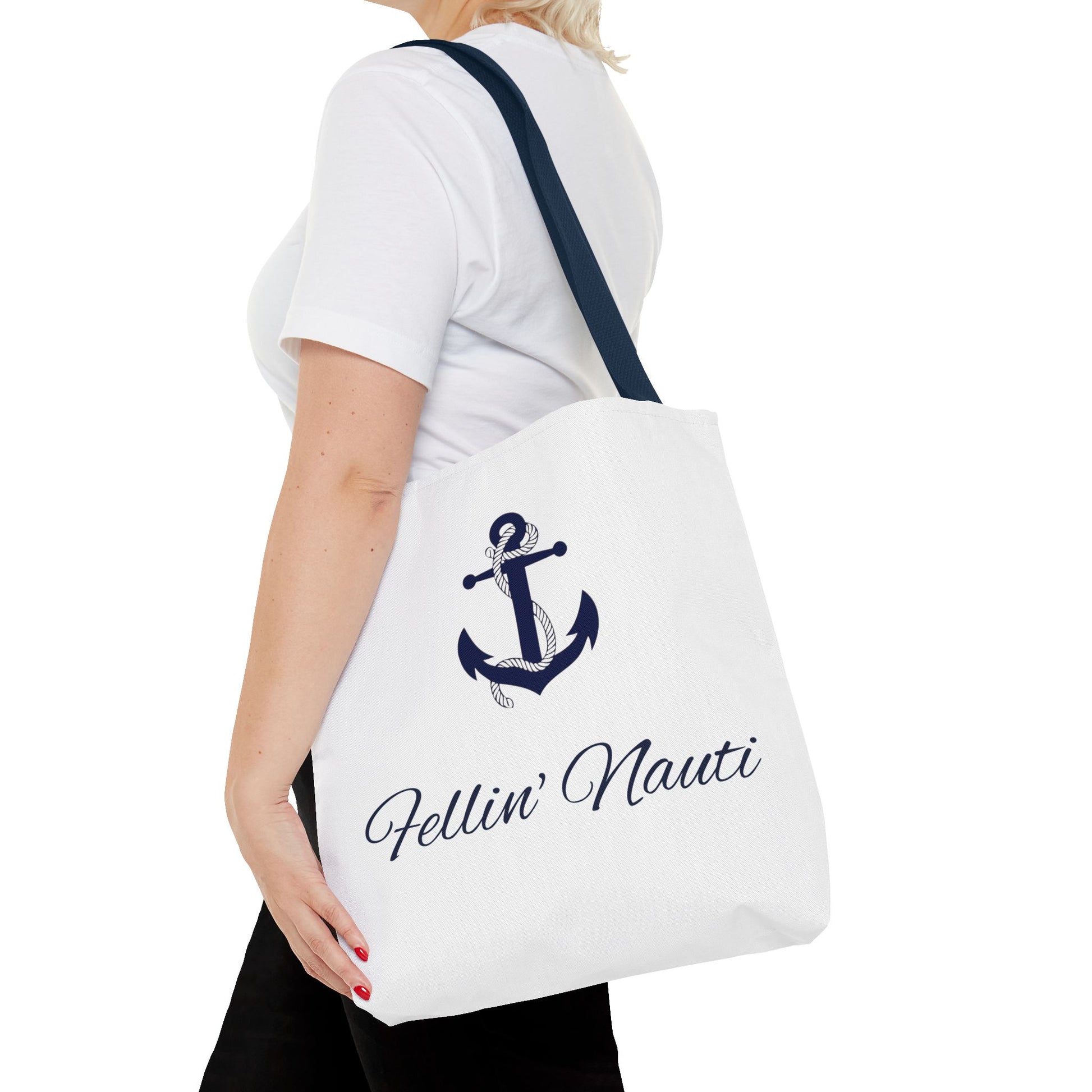A white canvas tote bag with a navy blue anchor graphic and the text 'Fellin' Nauti' in script, shown being worn by a model in a white t-shirt and black pants against a white background.