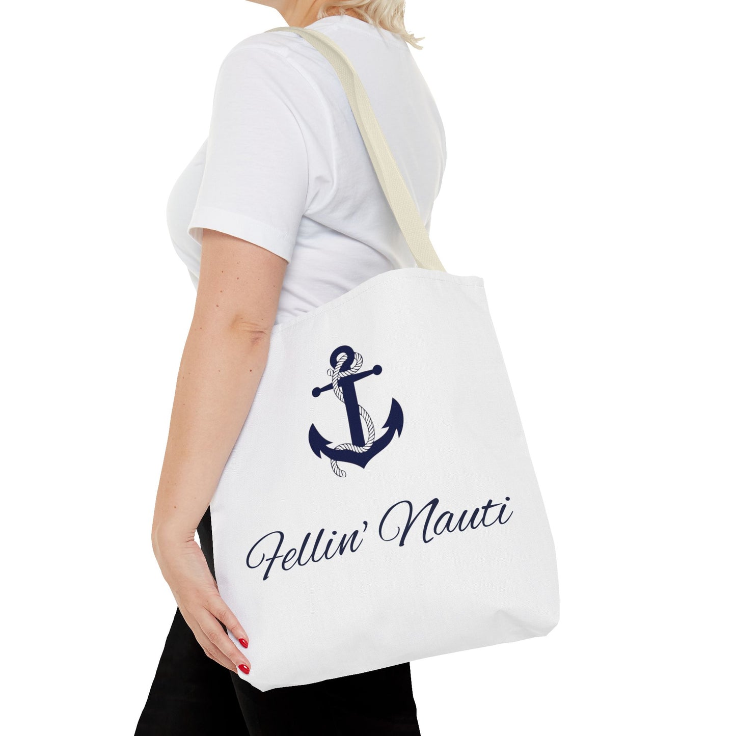 A white canvas tote bag with a navy blue anchor graphic and the text 'Fellin' Nauti' in cursive, shown being worn by a model in a white t-shirt and black pants against a white background.