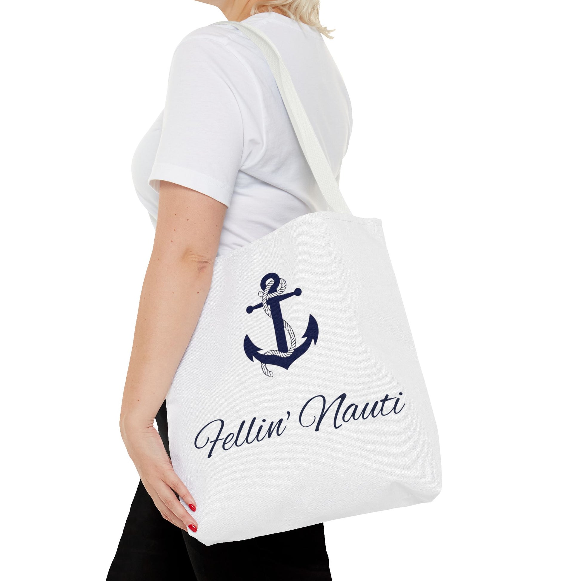 A white canvas tote bag with a navy blue anchor graphic and the text 'Fellin' Nauti' in cursive, shown being worn by a model in a white t-shirt and black pants against a white background.