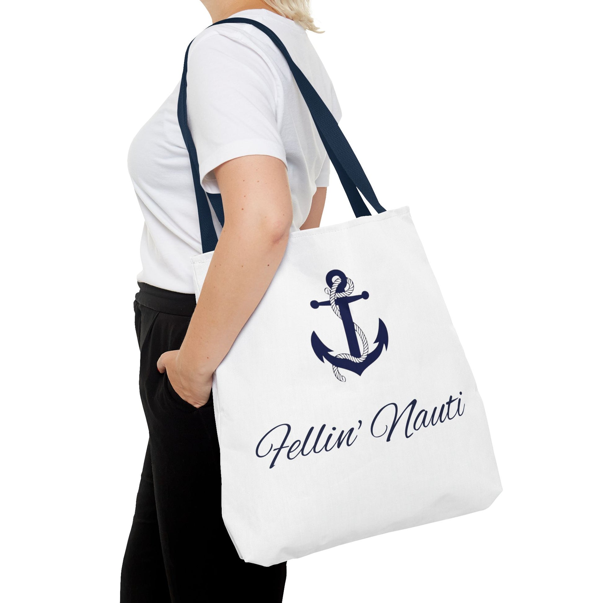 A white canvas tote bag with dark navy blue straps and a navy blue anchor graphic, featuring the text 'Fellin' Nauti' in a cursive font. The bag is shown being worn by a model wearing a white t-shirt and black pants against a plain white background.