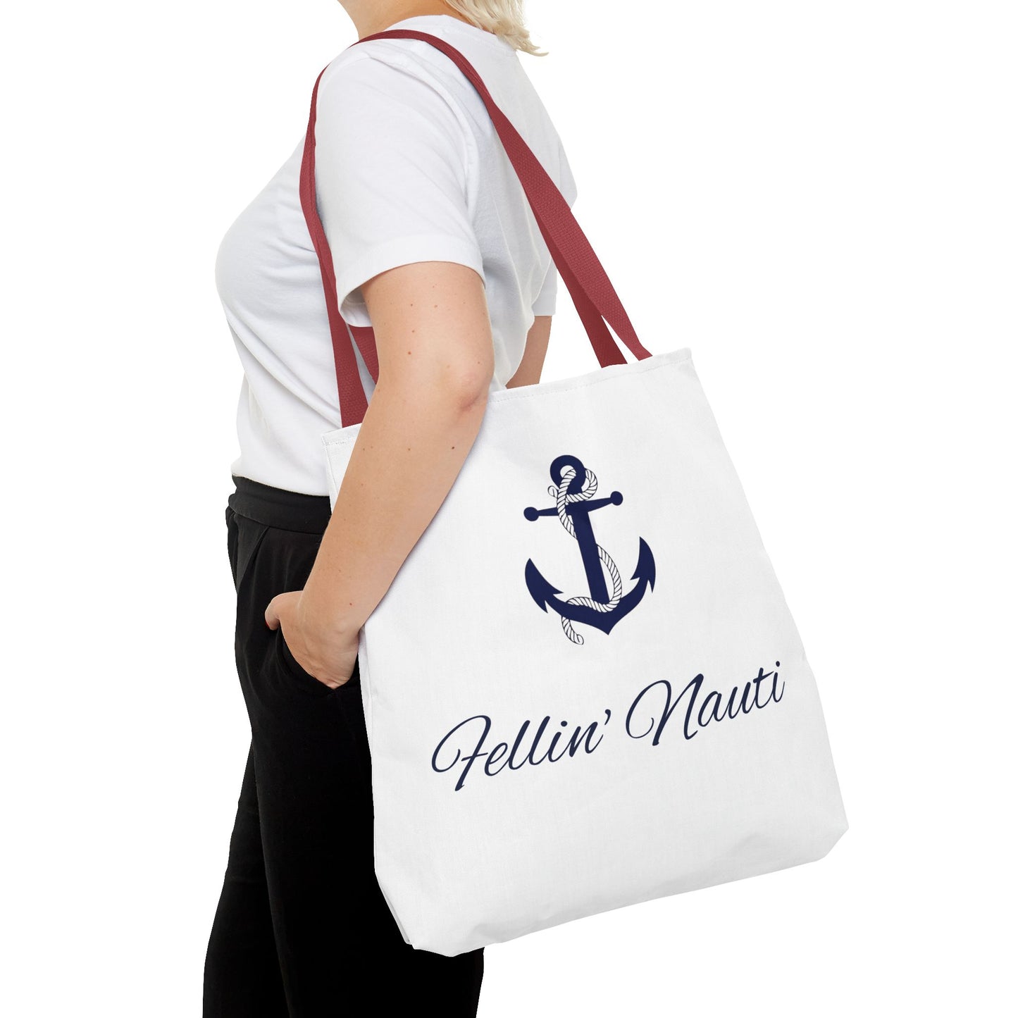 A white canvas tote bag with a navy blue anchor graphic and the text 'Fellin' Nauti' in navy script, carried on the shoulder of a person wearing a white t-shirt and black pants against a white background.