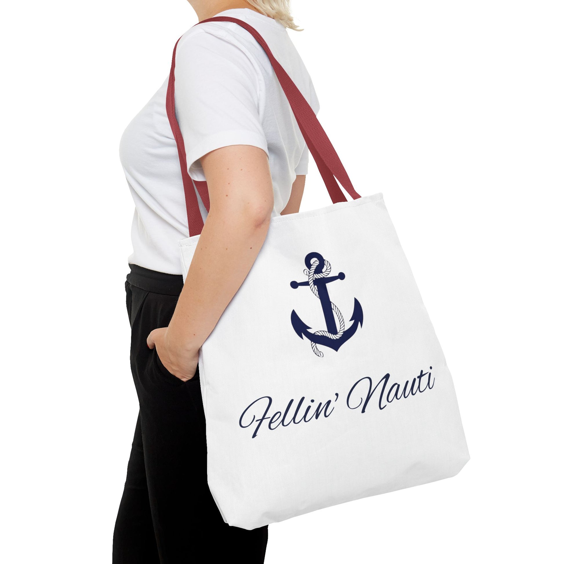 A white canvas tote bag with a navy blue anchor graphic and the text 'Fellin' Nauti' in navy script, carried on the shoulder of a person wearing a white t-shirt and black pants against a white background.