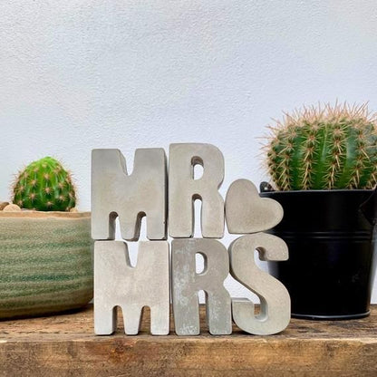 Decorative sign with 'MR & MRS' letters between two cacti on a wooden surface.