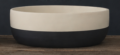 A two-tone cement bowl with a matte cream upper half and a dark charcoal black lower half, resting on a rustic wooden surface against a dark background.