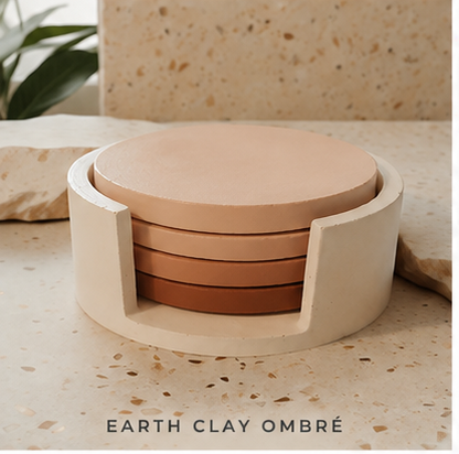 A set of round cement coasters in an earth clay ombré color gradient, housed in a matching cylindrical holder, displayed on a textured stone surface with a plant in the background.