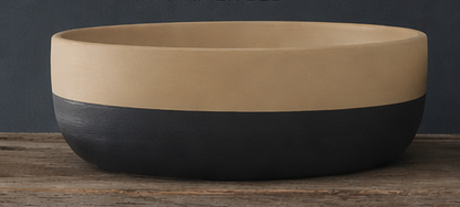 A two-tone cement bowl with a matte beige upper half and a dark charcoal black lower half, displayed on a rustic wooden surface against a dark gray background.