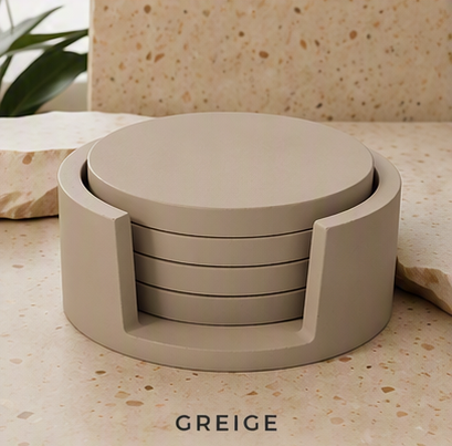 A round set of four cement coasters in a matte greige color, shown in their storage holder on a speckled stone surface with a plant in the background. The word 'GREIGE' is printed at the bottom.