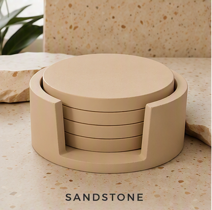 A set of round cement coasters in a matte sandstone finish, displayed in a cylindrical holder on a speckled stone surface with a matching stone background. The word 'SANDSTONE' is printed at the bottom.