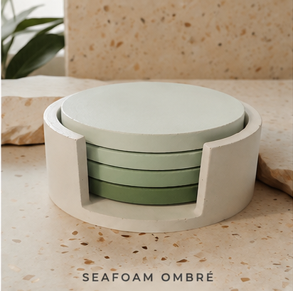 A set of round cement  coasters in a seafoam ombré gradient, housed in a matching cylindrical holder, displayed on a speckled beige stone surface with a plant in the background.