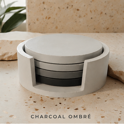 A round cement  coasters set in a matte finish, featuring a charcoal ombré color gradient from light gray to black. The set is displayed on a speckled beige stone surface with a matching stone background and a green plant leaf in the corner.