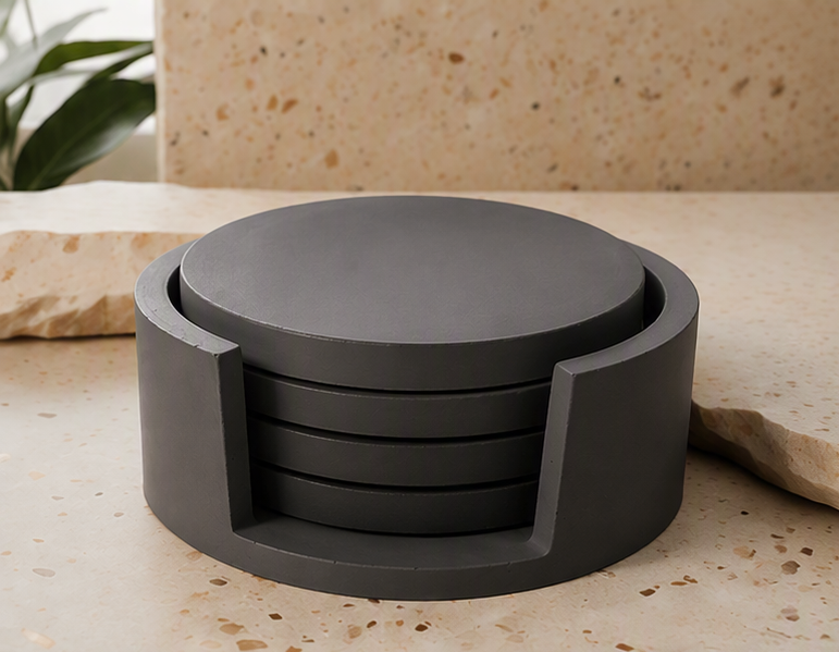 A dark charcoal gray circular cement coaster set with a matching top coaster, displayed on a beige stone surface with a matching stone background.