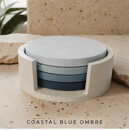 A set of four cement  round coasters in a blue ombré gradient, housed in a matching white cylindrical holder, displayed on a beige terrazzo surface with the text 'COASTAL BLUE OMBRÉ' at the bottom.