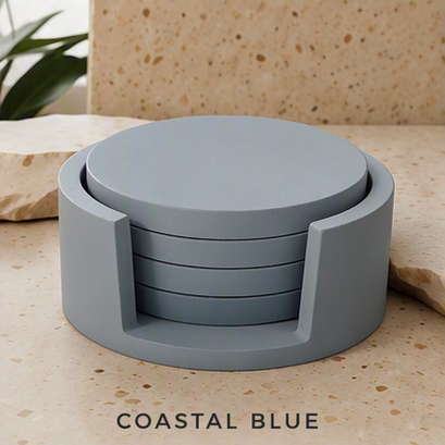 A set of round cement coasters in a muted coastal blue-gray color, displayed on a speckled beige stone surface with a plant and stone slab in the background. The text 'COASTAL BLUE' is printed at the bottom.