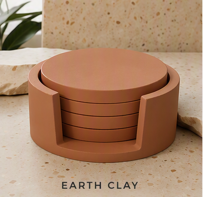 A set of four round cement coasters in a matte earth clay terracotta color, nested inside a matching cylindrical holder. The coasters and holder are displayed on a speckled beige stone surface with a matching stone background and a green plant leaf in the upper left corner.