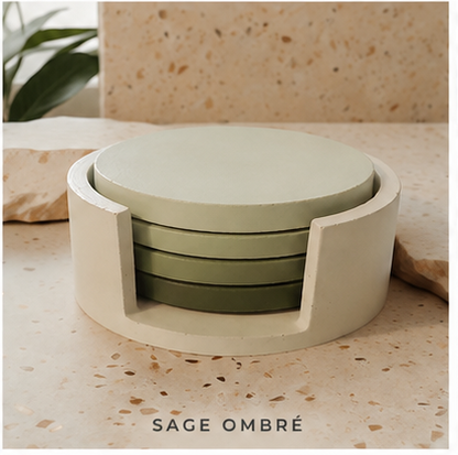 A set of four round  cement coasters in a sage green ombré color, housed in a matching cream-colored cylindrical holder, displayed on a speckled beige stone surface with a plant leaf in the background.