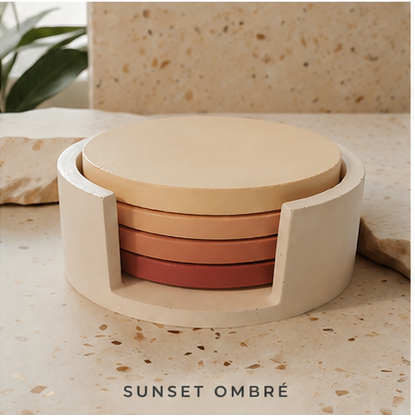 A set of four round cement coasters stacked inside a matching round holder, shown on a stone surface with a stone block and plant in the background. The coasters have a warm gradient from cream to terracotta, labeled 'SUNSET OMBRE' at the bottom.